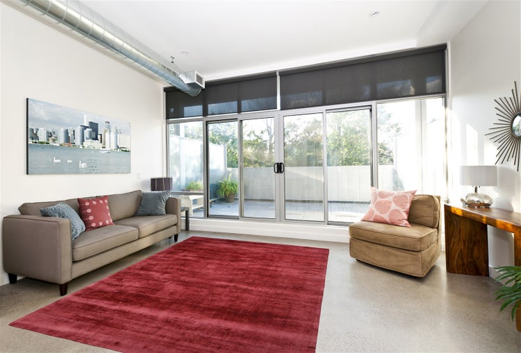 Helles Wohnzimmer mit grauem Sofa, rotem Teppich, Glasschiebetüren und Rollos. Sichtbares Lüftungsrohr aus Metall und moderner Beistelltisch.