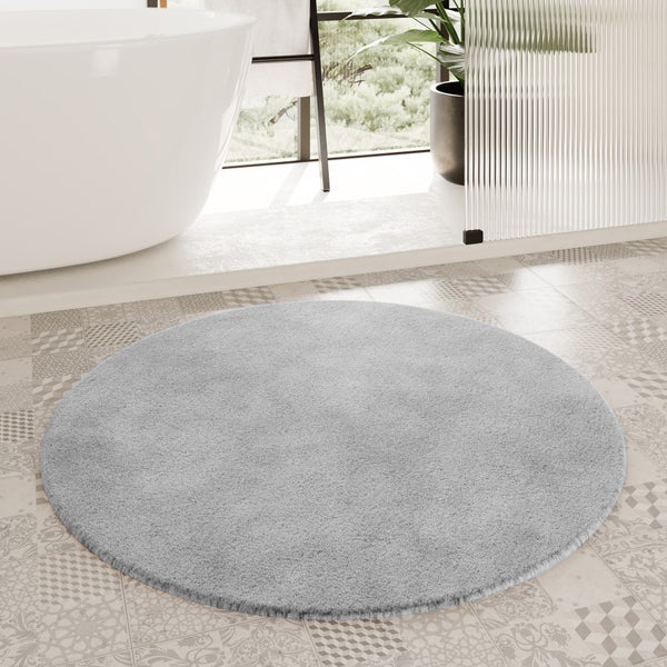 Runder, hellgrauer Badeteppich aus weichem Plüsch in einem hellen Badezimmer mit Badewanne und gemusterten Fliesen.