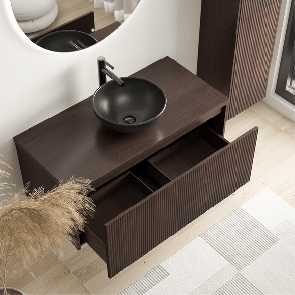 Moderner Waschbeckenunterschrank in dunkler Holzoptik mit geriffelter Front, einer offenen Schublade, schwarzem Aufsatzbecken und Armatur.