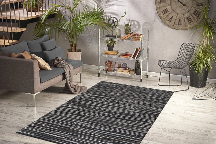 Heller, organisierter Wohnbereich mit grauem Sofa, schwarz-grau gestreiftem Teppich, Metallregal, schwarzem Drahtstuhl und Zimmerpflanzen.