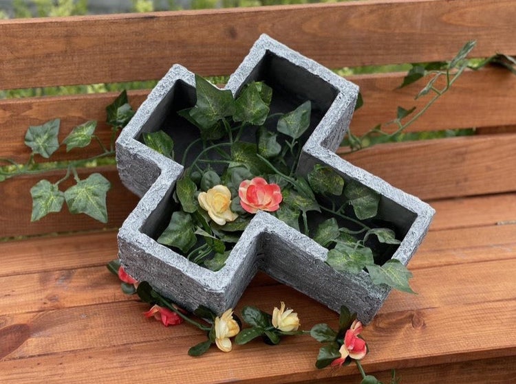 Grabschale in Kreuzform als Pflanzgefäß aus Steinoptik, dekoriert mit künstlichen Efeuranken und Blumen für die Grabgestaltung.