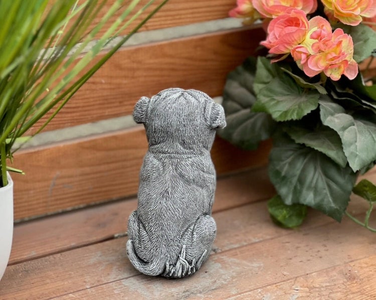 Gartenfigur sitzender Hund aus Stein von hinten, dekorative Steinfigur auf Holzoberfläche zwischen Pflanzen.