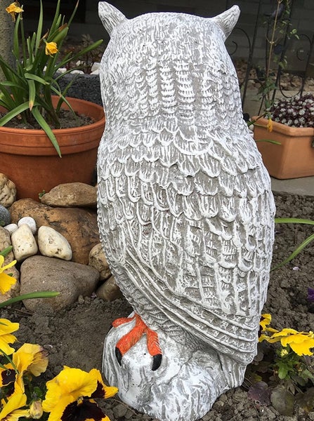 Detaillierte Eulen Gartenfigur aus Steinguss mit strukturierter Oberfläche, platziert in einem Gartenbeet zwischen Blumen und Steinen.