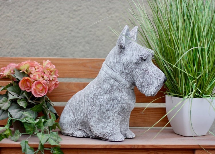 Steinfigur in Form eines sitzenden Scottish Terriers auf einer Holzbank, flankiert von Topfblumen und Ziergras.