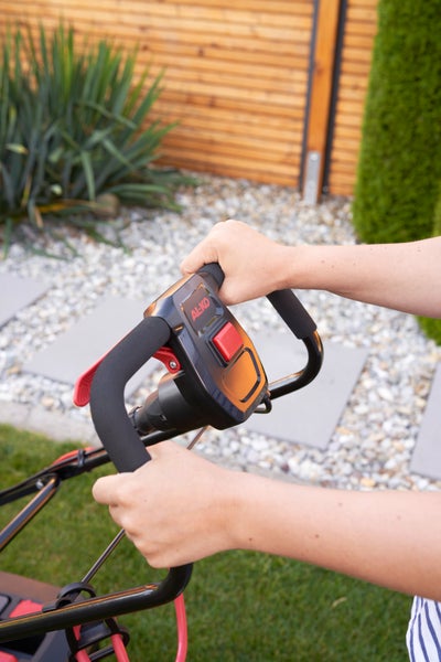 Hände an der ergonomischen Griffstange eines AL-KO Rasenmähers mit Sicherheitsbügel und rotem Startknopf in einem Garten
