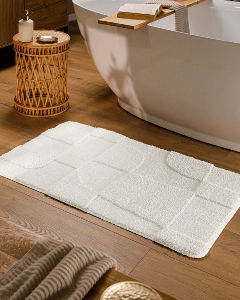 Weißer Badeteppich mit geometrischem Muster auf Holzboden neben einer weißen Badewanne und einem Korbtisch in einem hellen Badezimmer.
