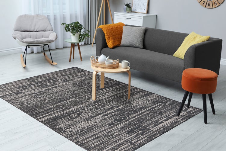 Modernes Wohnzimmer mit großem, grau-weiß gemustertem Teppich, dunklem Sofa, Schaukelstuhl und Holztisch auf hellem Boden.