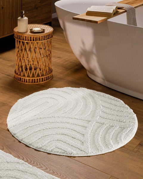 Runder weißer Badeteppich mit Wellenstruktur auf Holzboden in einem modernen Badezimmer neben einer Badewanne und einem Rattantisch.
