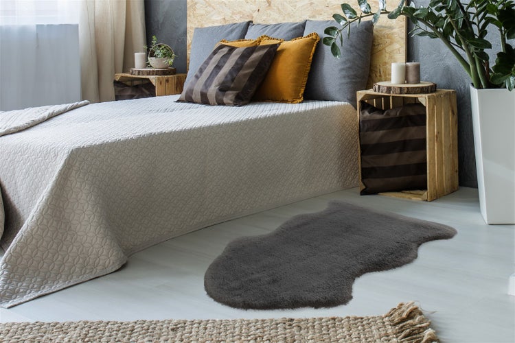 Grauer Kunstfell-Teppich in einem hellen Schlafzimmer mit beiger Tagesdecke, Nachttischen aus Holzkisten und Dekokissen.