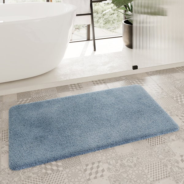 Hellblaue, flauschige Badematte auf gemusterten Fliesen neben einer weißen freistehenden Badewanne in einem hellen Badezimmer.