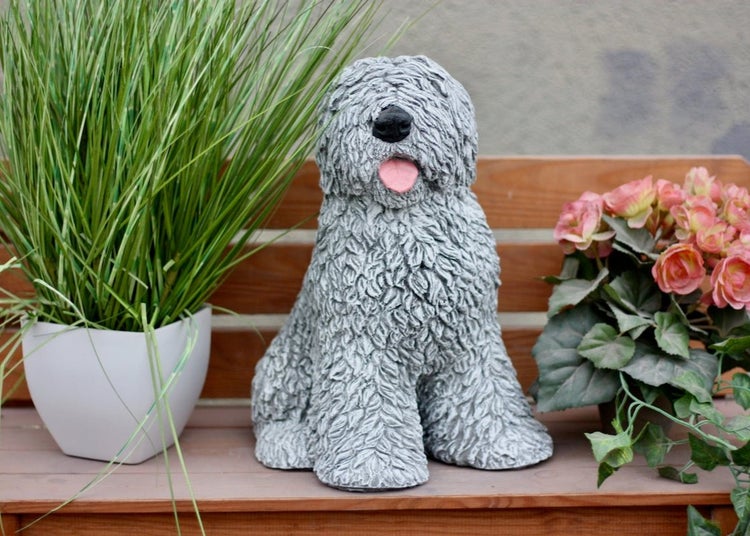 Gartenfigur in Form eines zotteligen Hundes, sitzend auf einer Holzbank zwischen Topfpflanzen.
