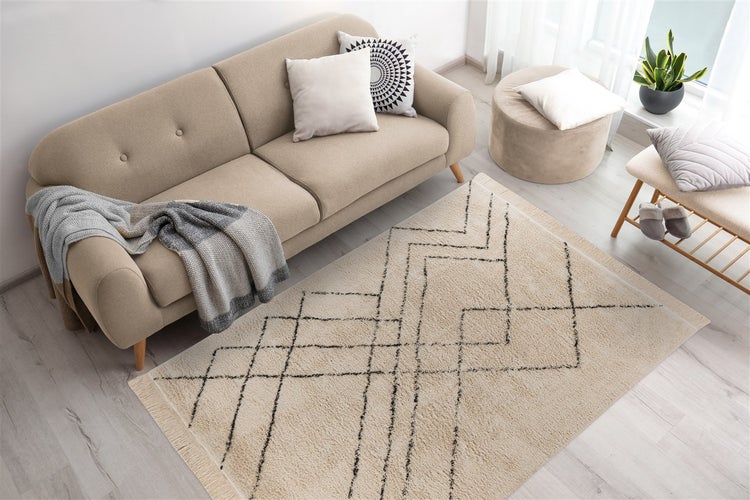 Helles Wohnzimmer mit beigefarbenem Sofa, Teppich mit schwarzem geometrischem Muster, Pouf und Holzbank auf hellem Holzboden.