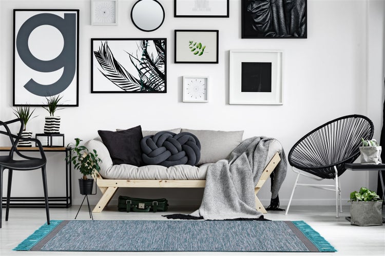 Moderner, heller Wohnraum mit blauem Webteppich mit Fransen, hellem Holzsofa mit grauen Kissen und einer Bilderwand mit verschiedenen Rahmen.