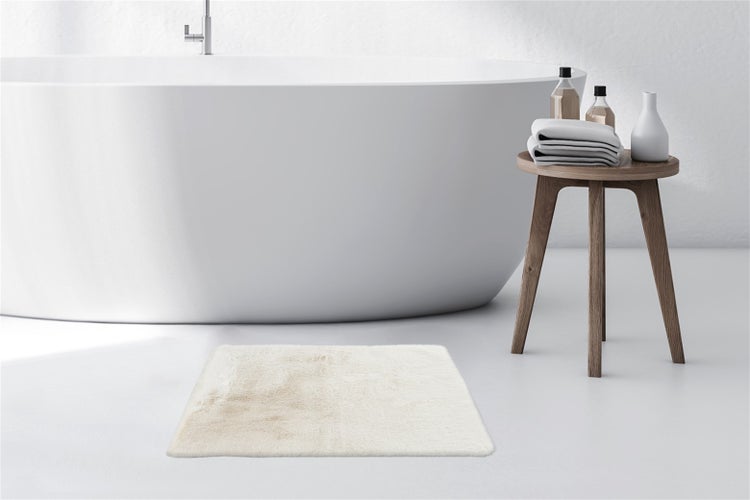 Rechteckige flauschige Badematte in einem hellen modernen Badezimmer vor einer freistehenden weißen Badewanne und einem Holzhocker mit Handtüchern.