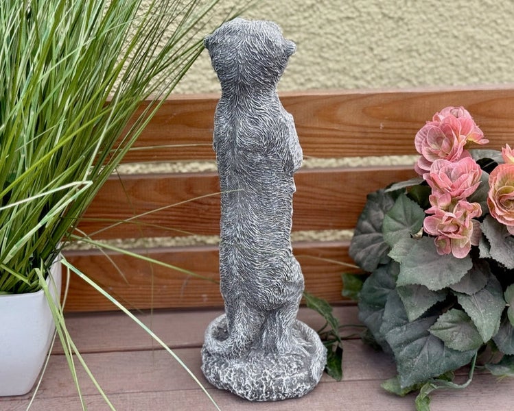 Steinfigur Erdmännchen in aufrechter Haltung als dekorative Gartenfigur auf einer Terrasse zwischen Pflanzen.