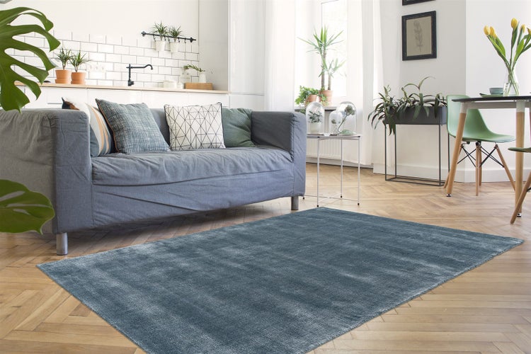 Blauer rechteckiger Teppich in modernem, hellem Wohnzimmer mit Parkettboden, grauem Sofa und dekorativen Zimmerpflanzen.