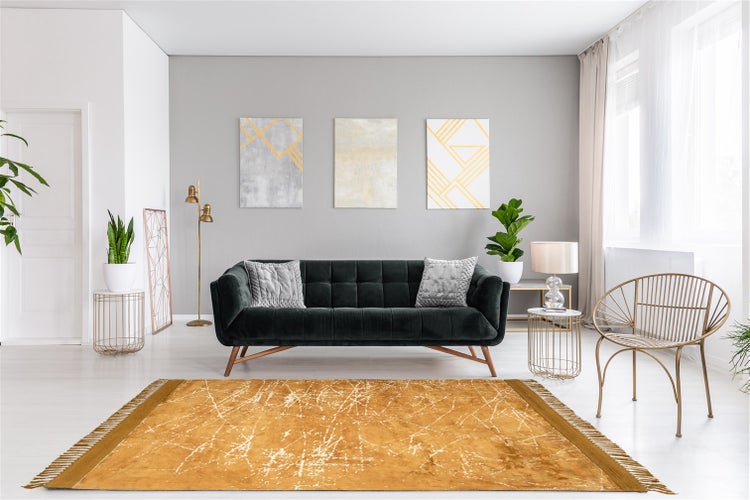 Modernes Wohnzimmer mit dunkelgrünem Sofa, senfgelbem Teppich mit Fransen, goldfarbenem Stuhl und abstrakten Wandbildern an einer grauen Wand.