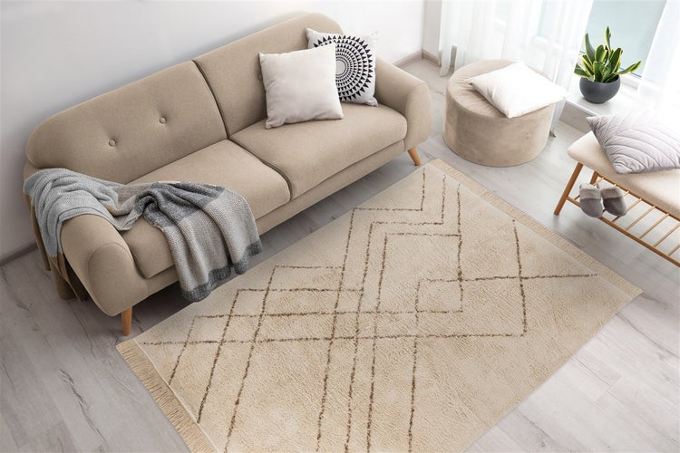 Beiger Teppich mit geometrischem Muster und Fransen in einem hellen Wohnzimmer mit Sofa, Pouf und Holzfußboden.
