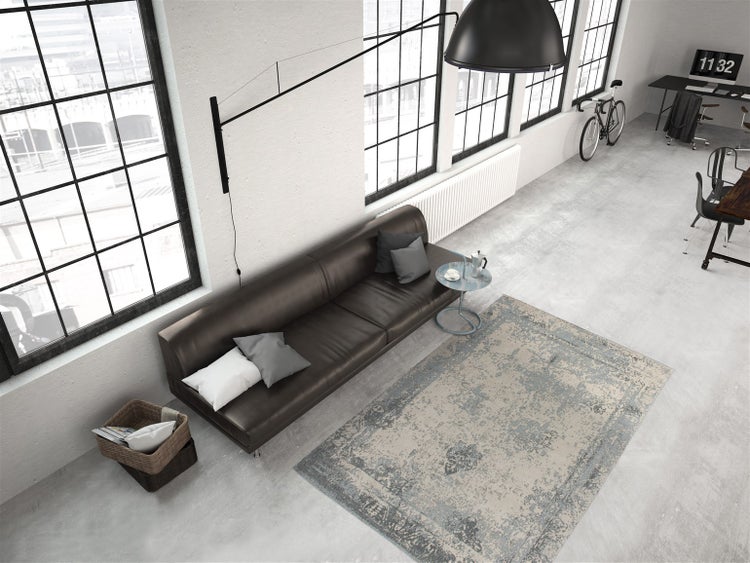 Großzügiges industrielles Loft mit großen Fenstern und einem grau-beigen Teppich im Vintage-Stil auf Betonboden neben einem braunen Ledersofa.