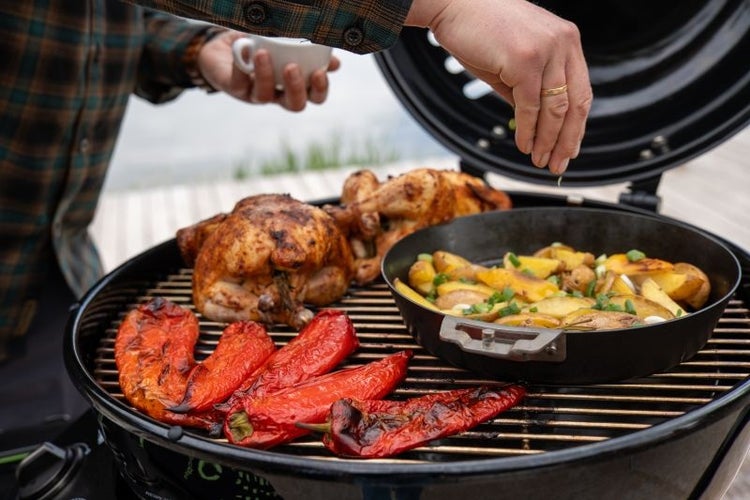 Ein runder Grill mit zwei ganzen Hähnchen, roten Paprika und einer gusseisernen Pfanne mit Kartoffeln, die mit Kräutern bestreut werden.