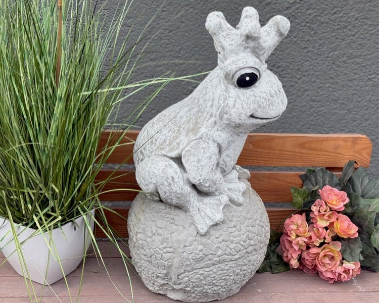 Gartenfigur Froschkönig aus Stein, sitzend auf einer Kugel, dekoriert auf einer Holzbank neben einer Pflanze und Blumen.
