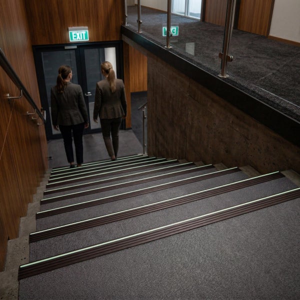 Treppenhaus mit Treppenkantenprofilen mit nachleuchtendem Streifen, Blick von oben auf die Treppe mit Holzwand und Ausgangsschild.