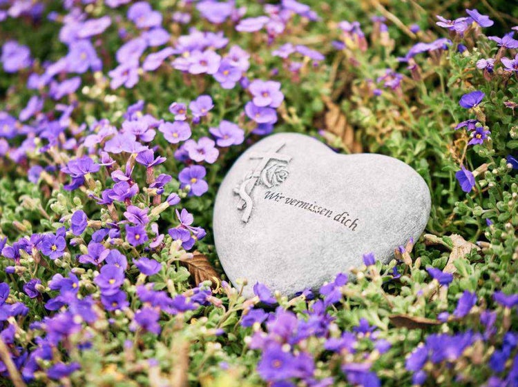 Herzfoermiger Gedenkstein aus Stein mit der Inschrift Wir vermissen dich und einem eingravierten Kreuz mit Rose zwischen violetten Blueten