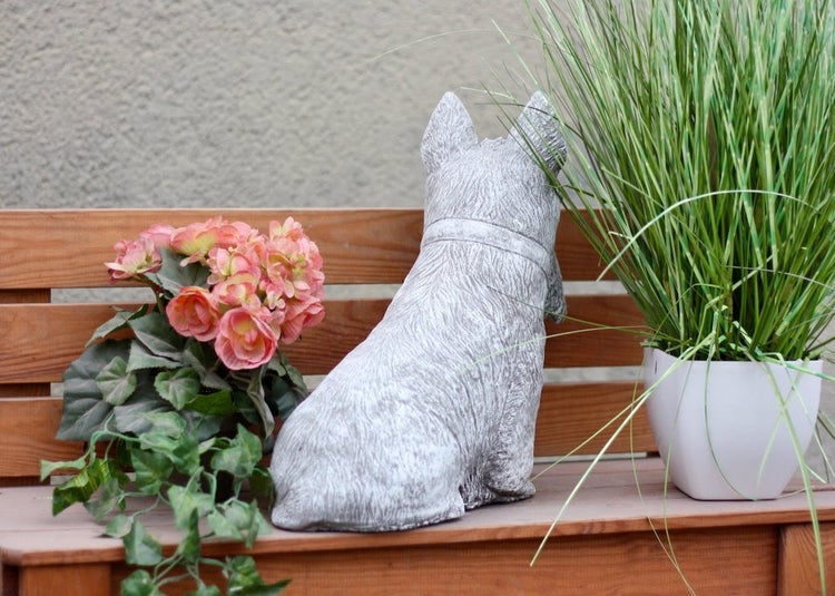 Gartenfigur in Form eines sitzenden Hundes aus Steinguss, von hinten fotografiert, auf einer Holzbank zwischen zwei Topfpflanzen.
