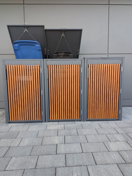 Dreier-Mülltonnenbox aus Metall mit vertikalen Holzlatten, zwei Deckel sind geöffnet.