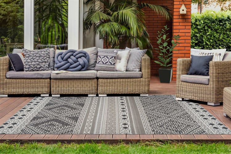 Moderner Außenteppich mit geometrischem Muster auf einer Holzterrasse vor einem Polyrattan-Sofa mit Dekokissen und Gartenpflanzen.
