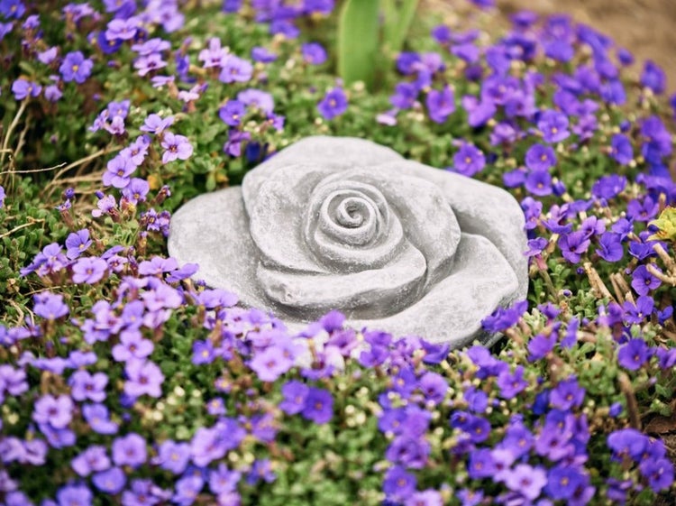 Dekorative Gartenfigur in Form einer Rose aus Stein inmitten von violetten Blumen im Garten