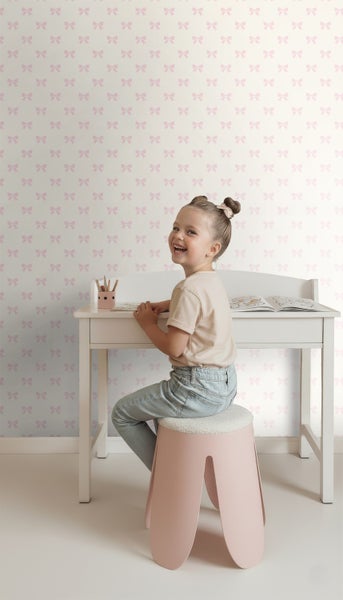 Helles Kinderzimmer mit Tapete im rosa Schleifenmuster. Ein Mädchen sitzt auf einem rosa Hocker an einem weißen Holzschreibtisch.
