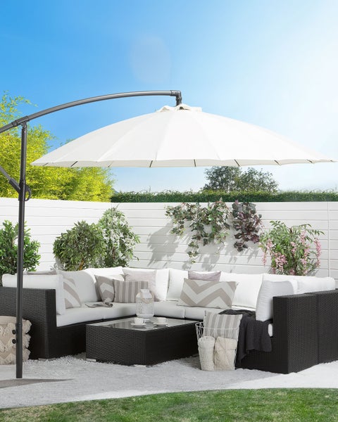 Moderne Gartenlounge aus dunklem Polyrattan mit hellen Polstern unter einem weißen Ampelschirm auf einer hellen Terrasse mit Pflanzen.