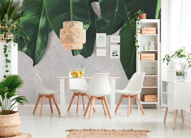 Vliestapete mit großflächigem Motiv grüner Bananenblätter in einem hellen Esszimmer mit weißem Tisch, Holzstühlen und geflochtener Hängeleuchte.