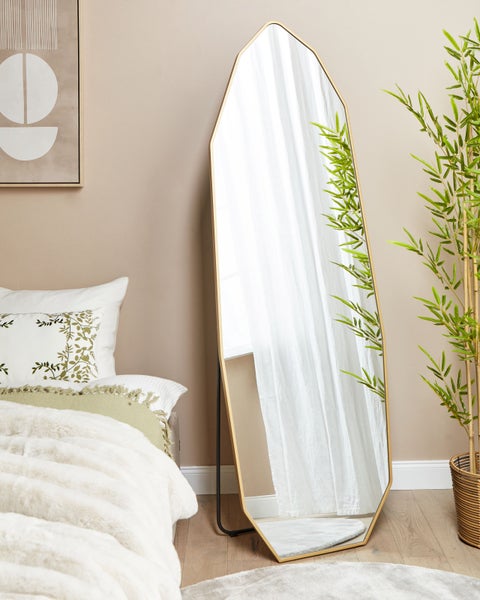 Dekorativer Standspiegel mit goldfarbenem Rahmen neben einem Bett und einer Zimmerpflanze.