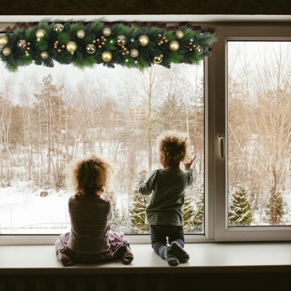 Zwei Kinder sitzen am Fenster mit Blick auf eine Winterlandschaft, geschmückt mit einer Tannengirlande und goldenen Kugeln.