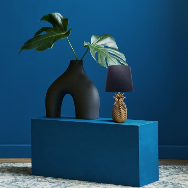 Dekorative Szene mit einer schwarzen Vase mit Monstera-Blättern und einer goldenen Ananaslampe auf einem blauen Podest.