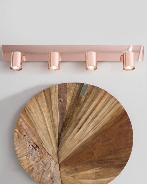 Moderne 4-flammige Strahler-Leiste aus kupferfarbenem Metall über einer runden Wanddekoration aus Holz mit radialer Struktur.