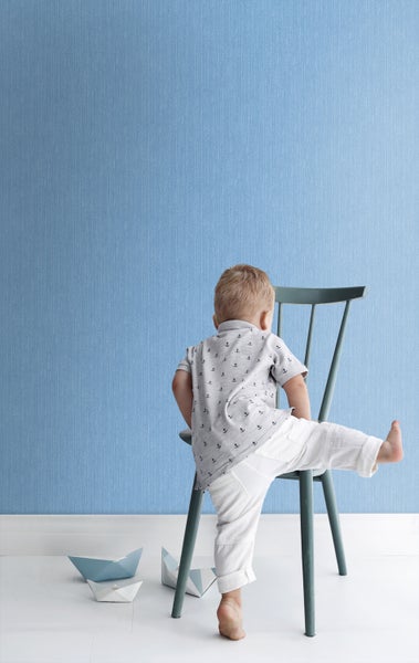 Ein Kind sitzt auf einem Stuhl vor einer blauen Tapete mit Papierbooten auf dem Boden.