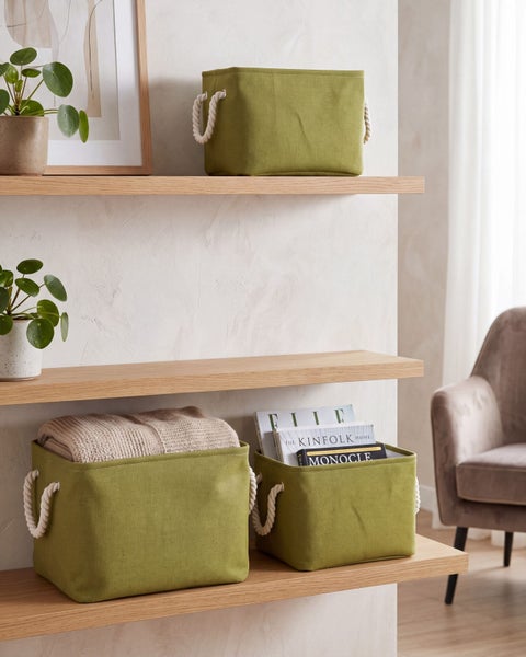 Grüne Aufbewahrungskörbe aus Stoff mit Seilgriffen auf Holzregalen, gefüllt mit einer Decke und Zeitschriften in einem hellen, geordneten Raum.