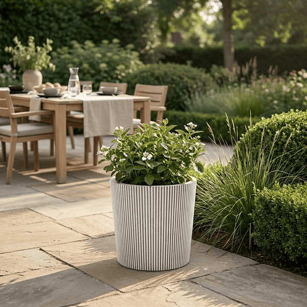 Geriffelter Pflanzkübel mit Pflanze auf einer Steinterrasse, im Hintergrund Holztisch und Stühle in einer hellen Gartenanlage.