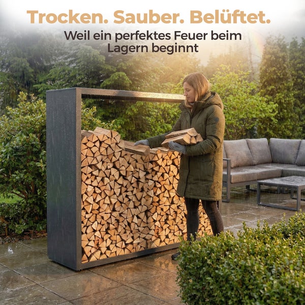 Moderne Szene auf einer Terrasse: Eine Frau stapelt Holzscheite in ein großes, anthrazitfarbenes Kaminholzregal aus Metall bei Regen.