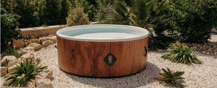 Runder Whirlpool in Holzoptik mit weißer Innenseite in einem hellen Garten mit Kieselsteinen und Pflanzen.