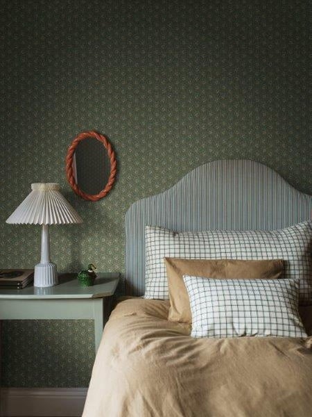 Schlafzimmer mit Bett, Nachttisch, Lampe und Spiegel vor gemusterter Tapete.