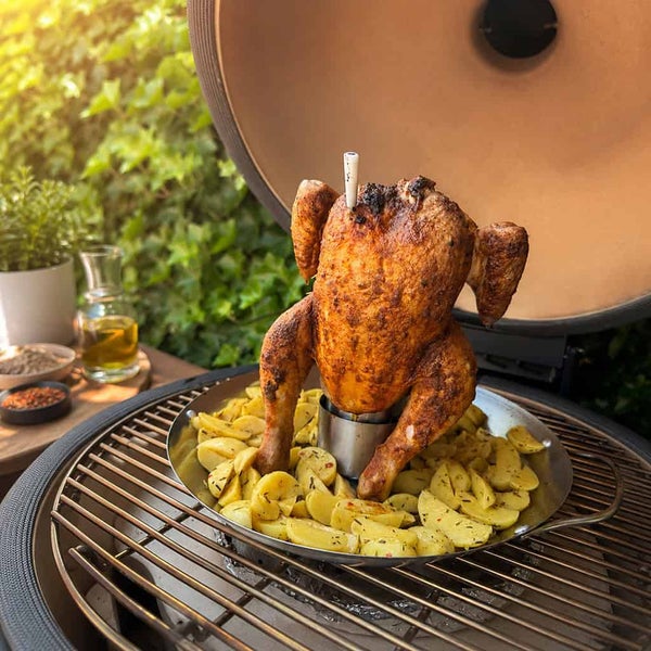 Ein ganzes gegrilltes Hähnchen auf einem Hähnchenbräter aus Edelstahl in einer Pfanne mit Kartoffelspalten auf einem Keramikgrill.