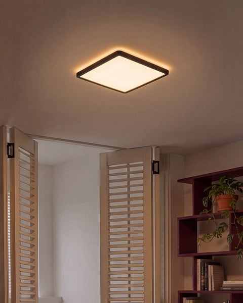 Quadratische LED Deckenleuchte mit flachem Design, montiert an einer Zimmerdecke über offenen Fensterläden und einem Regal mit Zimmerpflanze.