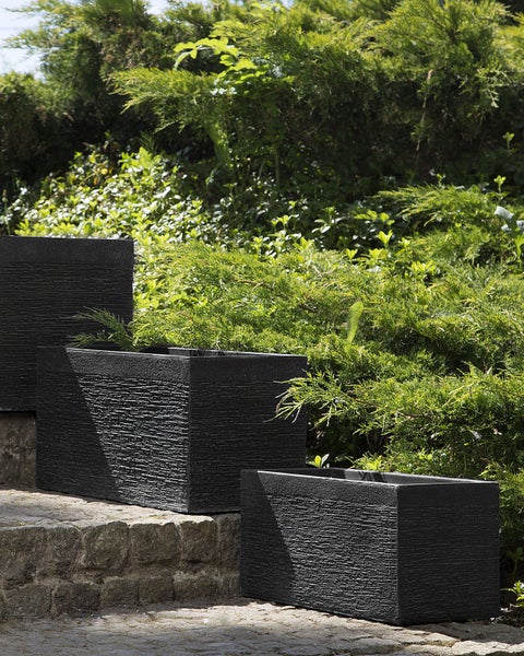 Drei dunkle Pflanzkästen aus Kunststoff mit Holzstruktur auf Steinstufen vor Grünpflanzen