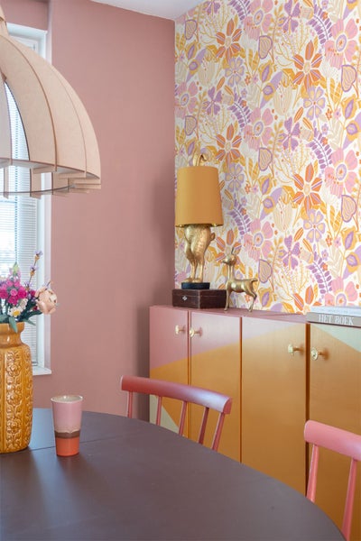 Ein helles Zimmer mit floraler Tapete, einem Sideboard mit geometrischem Farbmuster, einer Hasen-Tischlampe und einem Esstisch mit Stühlen.