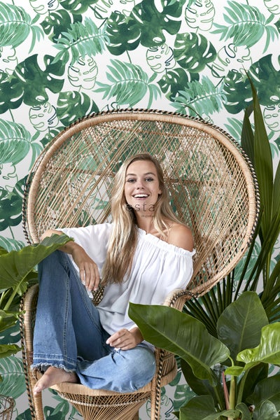 Frau sitzt in einem Rattan-Pfauenstuhl vor einer botanischen Tapete