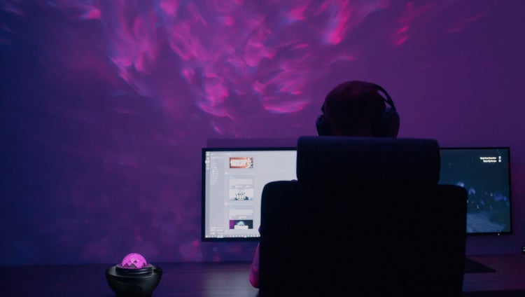 Szene mit einem Zimmer, beleuchtet mit einem Sternenhimmel-Projektor und einem Mann, der Kopfhörer trägt und vor zwei Computermonitoren sitzt